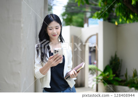 Young woman leaning against wall, using phone while holding notebook outside a cozy cafe Young woman leaning against wall, using phone while holding notebook outside a cozy cafe 128408944