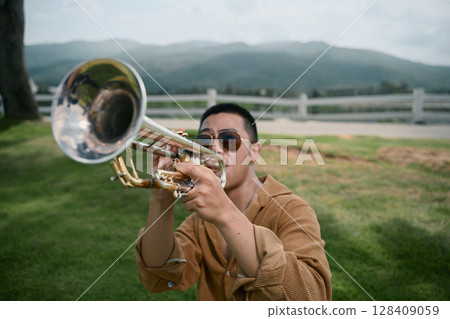Artistic outdoor shot of man playing trumpet outdoors with mountain scenery in the background 128409059