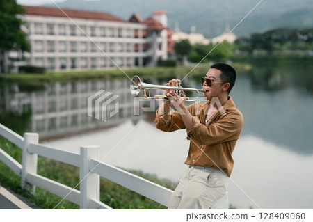 Man playing trumpet near a serene lake with building and mountains in the background Man playing trumpet near a serene lake with building and mountains in the background 128409060