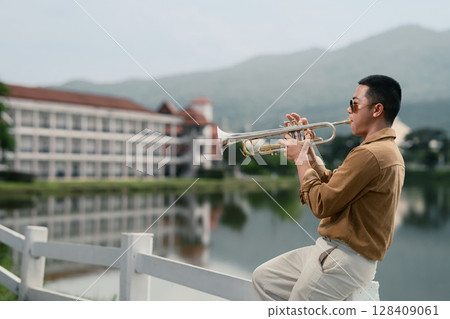 Man playing trumpet near a serene lake with building and mountains in the background 128409061