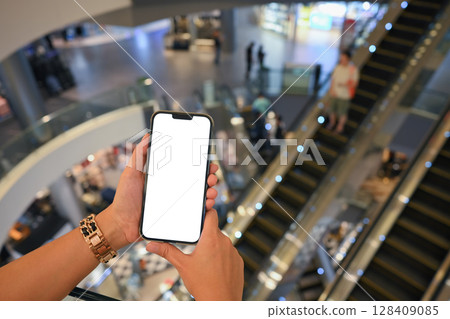 Hands holding smartphone with blank screen inside a modern shopping mall 128409085