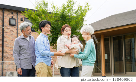 A three-generation family smiling and surrounding a baby in front of their house 128409863