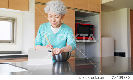 一位老年婦女一邊做飯一邊看平板電腦 一位老年婦女一邊做飯一邊看平板電腦 128409870