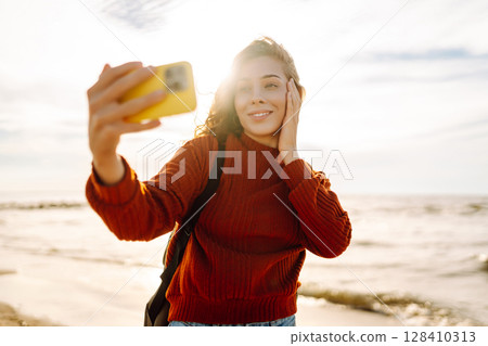 A joyful young woman takes a selfie on a sunny beach at sunset while enjoying the ocean waves. 128410313