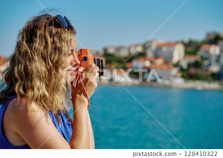 Woman standing near sea holding instant camera and taking photo. Female tourist at coastal resort with blurred town in background. Travel and summer vacation concept Woman standing near sea holding instant camera and taking photo. Female tourist at coastal resort with blurred town in background. Travel and summer vacation concept 128410322