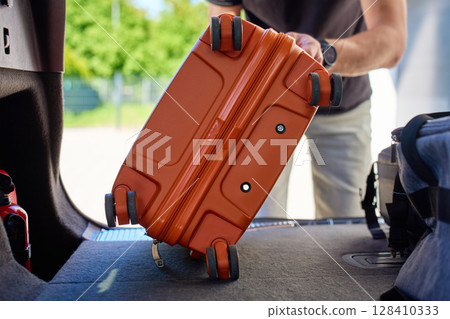 Close up of hand placing orange suitcase into car trunk, preparing for travel or vacation. Inside view of man loading luggage in vehicle 128410333