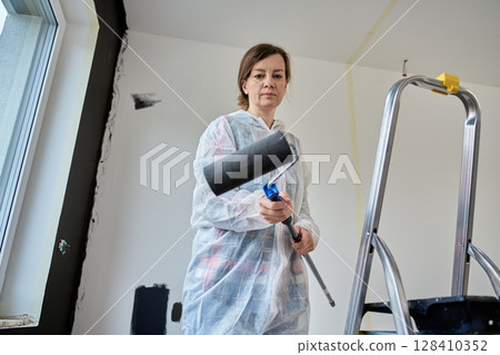 Woman in protective suit holds paint roller while preparing to paint white wall during interior renovation. Concept of self-made home improvement and budget repair Woman in protective suit holds paint roller while preparing to paint white wall during interior renovation. Concept of self-made home improvement and budget repair 128410352