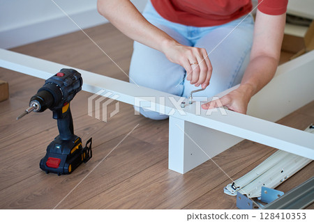 Woman assembling bed frame on wooden floor, using wrench and drill for installation. DIY furniture assembly and home improvement 128410353