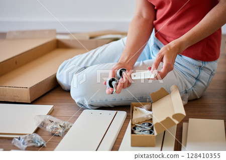 Woman sitting on floor assembling bed parts, holding wheels and hardware. Concept of DIY furniture assembly, home improvement and self installation process 128410355