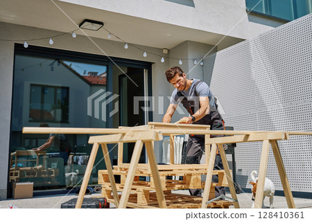 Man working on outdoor carpentry task, measuring wooden board on sawhorses in backyard Man working on outdoor carpentry task, measuring wooden board on sawhorses in backyard 128410361
