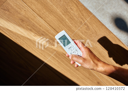 Close-up of person hand holding air conditioner remote control on sunny day with shadows. Adjusting temperature at home. Concept of climate control and cooling during hot weather Close-up of person hand holding air conditioner remote control on sunny day with shadows. Adjusting temperature at home. Concept of climate control and cooling during hot weather 128410372