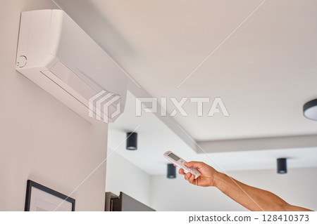 Close-up of hand holding remote control and adjusting temperature on wall mounted air conditioner at home. Concept of cooling and climate control during hot weather 128410373