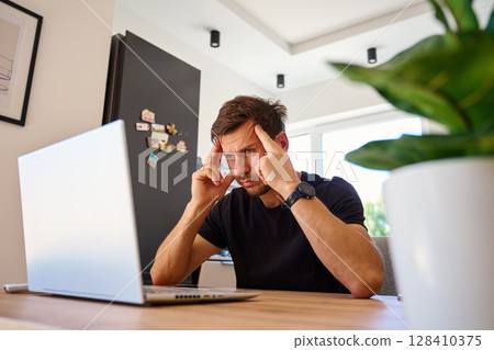 Exhausted person sitting at kitchen with face in hands. Stressed man working on laptop at home office desk. Concept of work pressure and burnout during remote work Exhausted person sitting at kitchen with face in hands. Stressed man working on laptop at home office desk. Concept of work pressure and burnout during remote work 128410375
