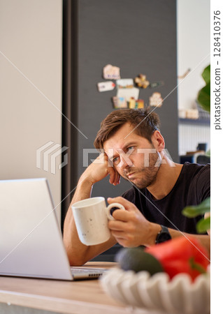 Tired man man working on laptop at home, holding coffee cup. Exhausted freelancer drinks coffee. Concept of remote work fatigue and burnout Tired man man working on laptop at home, holding coffee cup. Exhausted freelancer drinks coffee. Concept of remote work fatigue and burnout 128410376