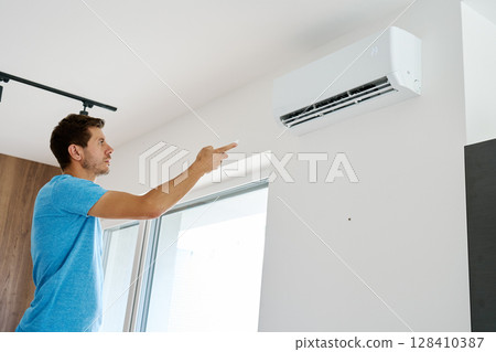 Man using remote control to adjust temperature on wall-mounted air conditioner in modern home interior. Concept of home cooling and indoor climate control during hot summer weather Man using remote control to adjust temperature on wall-mounted air conditioner in modern home interior. Concept of home cooling and indoor climate control during hot summer weather 128410387
