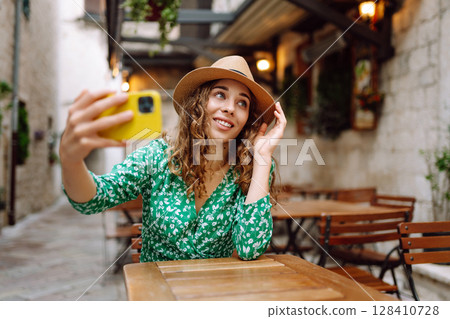 Young woman holding a smartphone in her hands and taking a selfie while sitting at the summer cafe. 128410728
