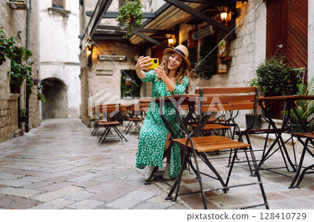 Young woman holding a smartphone in her hands and taking a selfie while sitting at the summer cafe. Young woman holding a smartphone in her hands and taking a selfie while sitting at the summer cafe. 128410729