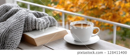 Cup of coffee beside an open book and candle on a window sill overlooking colorful autumn leaves, perfect for relaxation and reading 128411400