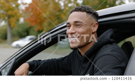 Smiling young man sits in the driver s seat of a car, surrounded by vibrant autumn trees and natural light 128411416