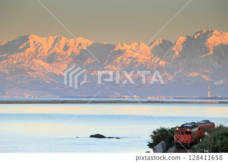 Himi Line Kiha 40 running along Amaharashi Coast with the Tateyama mountain range dyed red by the setting sun in the background 128411658