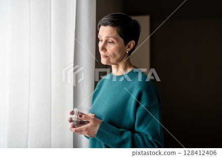 Exhausted woman with water medicine by window in stillness alone. Emotional tress, quiet reflection 128412406