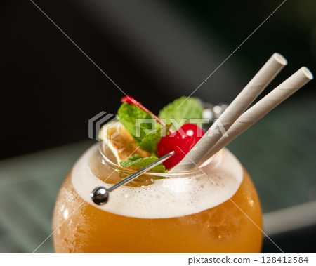 ibrant orange alcoholic cocktail, served in a unique round glass with a stem, is garnished with mint, berries, and two straws, all set on a bar counter 128412584