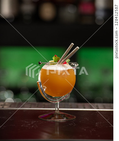 ibrant orange alcoholic cocktail, served in a unique round glass with a stem, is garnished with mint, berries, and two straws, all set on a bar counter 128412587