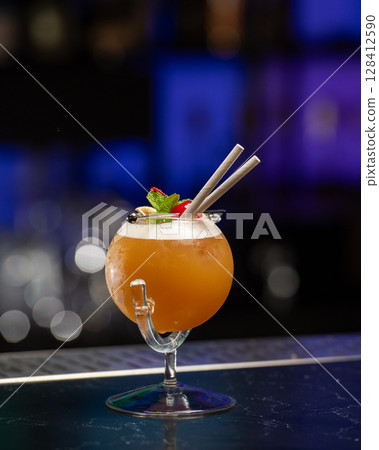 ibrant orange alcoholic cocktail, served in a unique round glass with a stem, is garnished with mint, berries, and two straws, all set on a bar counter 128412590