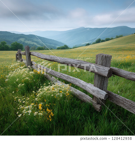 Scenic landscape featuring a weathered fence dividing a blooming meadow from rolling hills and distant mountains under a soft, overcast sky. Perfect for themes of nature, serenity, and rural beauty. 128412938