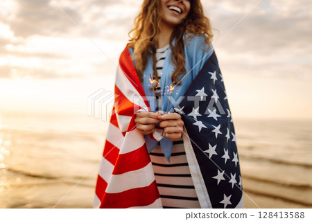 Young woman with American flag and sparklers on beach. Patriotic holiday. USA celebrate 4th of July. 128413588