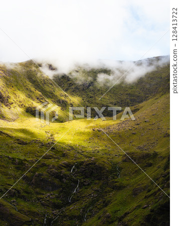 A valley within Carrauntoohil is covered in green vegetation, with sparse waterfalls cascading down the rock face and a heavy fog settling over the peaks in County Kerry. A valley within Carrauntoohil is covered in green vegetation, with sparse waterfalls cascading down the rock face and a heavy fog settling over the peaks in County Kerry. 128413722