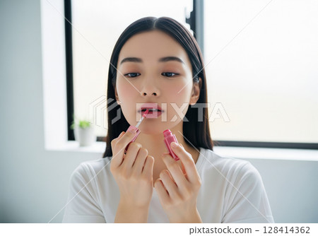 A woman applying pink lip gloss to her lips indoors with the soft morning light coming in through the window 128414362