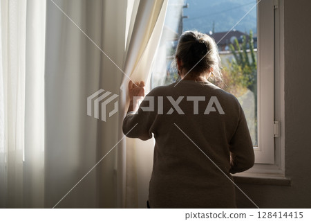 Lonely Elderly Woman Standing by the Window Looking Outside Waiting for a Visitor 128414415