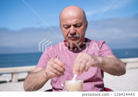 Elderly Man Adding a Lot of Sugar to Coffee While Sitting on a Summer Terrace Elderly Man Adding a Lot of Sugar to Coffee While Sitting on a Summer Terrace 128414443