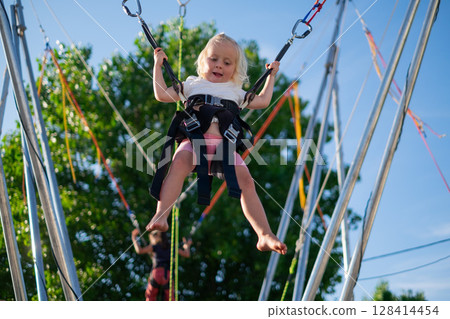Little European Girl Learning to Jump on a Trampoline with Safety Harness. Summer Fun Little European Girl Learning to Jump on a Trampoline with Safety Harness. Summer Fun 128414454