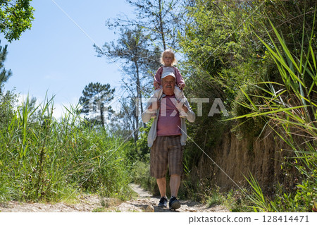 Grandfather Carrying His Granddaughter on His Shoulders Because She Got Tired During a Nature Hike 128414471