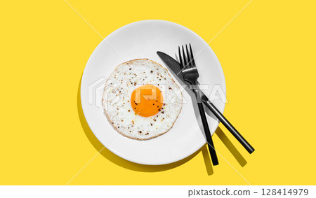 Fried eggs with spicy and black cutlery on a white plate on a bright yellow background with shadow. Fried eggs with spicy and black cutlery on a white plate on a bright yellow background with shadow. 128414979