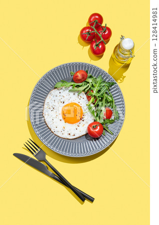Fried eggs with arugula and cherry tomatoes on a dark plate on a yellow background with cultery 128414981