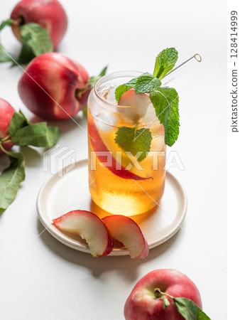 Ice tea with peach, mint in a glass on a white table near window with fresh fruits and sunlight. 128414999