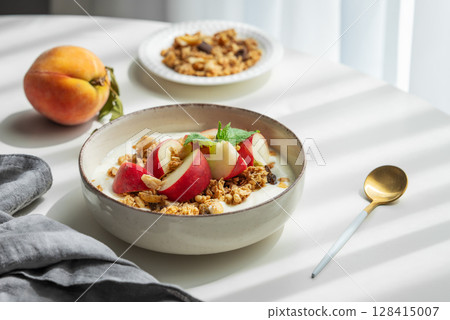 Natural greek yogurt with granola or muesli and peach in a bowl on a white table near window 128415007