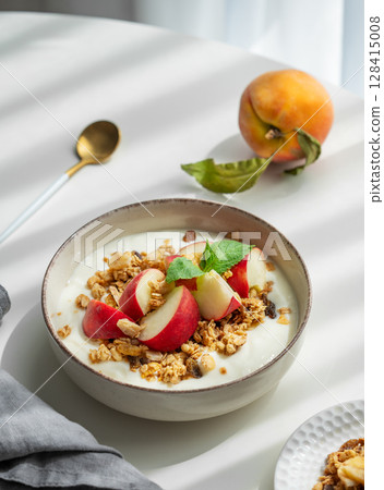 Natural greek yogurt with granola and peach in a bowl on a white table near window with spoon 128415008
