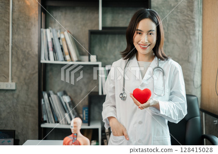 Female doctor in white coat holding heart model, standing in a modern medical office with books and anatomical model. 128415028