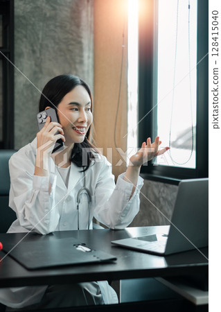 Female doctor in white coat talking on phone and using laptop in a bright, modern office. 128415040