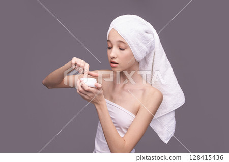 Young asian female applying skincare cream with towel on head against gray Young asian female applying skincare cream with towel on head against gray 128415436