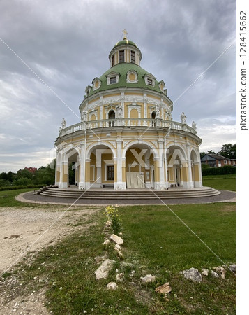 Baroque-style church with green dome framed by cloudy sky and vibrant greenery 128415662