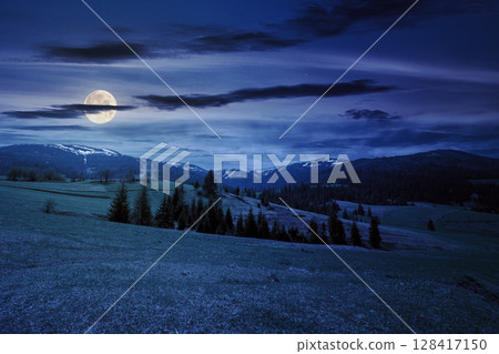 wide mountainous countryside landscape at night. forest on the rolling hills in the valley. distant ridge in full moon light under a dramatic starry sky with dark clouds 128417150