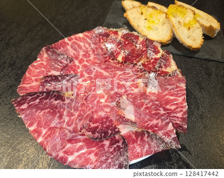 Gourmet cecina platter with artisan bread and olive oil on dark stone background. Cecina is the famous Spanish salted and dried meat. Made from Angus beef. Top view. Flat lay 128417442