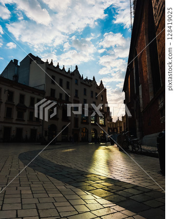 Sunlight reflecting off cobblestone street creates warm glow, highlighting historic buildings and St. Marys Basilica in Krakow, Poland, during picturesque sunset against cloudy blue sky. Historic city Sunlight reflecting off cobblestone street creates warm glow, highlighting historic buildings and St. Marys Basilica in Krakow, Poland, during picturesque sunset against cloudy blue sky. Historic city 128419025