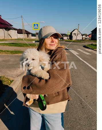 Smiling woman wearing casual clothing and cap holds small white poodle dog in her arms while standing in residential area near crosswalk on sunny day. Casual clad woman embracing white cute poodle 128419036