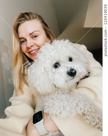Happy young woman wearing beige sweatshirt, smiling while holding her fluffy white dog, enjoying cozy moment at home filled with warmth and affection. Cheerful blonde woman embracing fluffy white dog 128419039